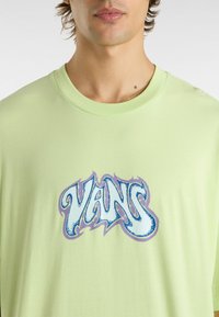 Camiseta de algodón verde claro con cuello redondo que presenta un gráfico en azul y morado del logo "VANS", sobre un fondo de textura suave.