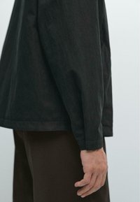 Black long-sleeve shirt with a relaxed fit, made of textured fabric; features simple cuffs and a smooth hem, worn over brown pants.