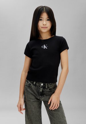 Dievča s dlhými tmavými vlasmi, ktoré má na sebe čierne tričko Calvin Klein a sivé džínsy, stojí pred jednofarebným svetlosivým pozadím.