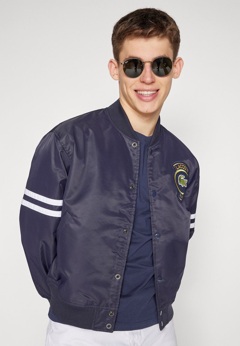 Veste bombardier marine en tissu lisse, avec des rayures blanches sur les manches et un logo brodé sur la poitrine.