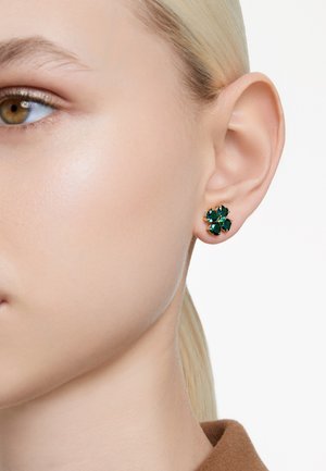 Zbliżenie na twarz kobiety, na której lewym uchu widoczny jest szmaragdowozielony kolczyk w kształcie czterolistnej koniczyny.