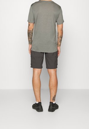 Hombre con camiseta gris, pantalones cortos cargo oscuros y zapatillas negras de pie de espaldas contra un fondo liso.