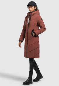 Kvinde iført en rustfarvet quilted knælang jakke med hætte, sorte bukser, sorte støvler og en sort baseballkasket, stående til venstre.