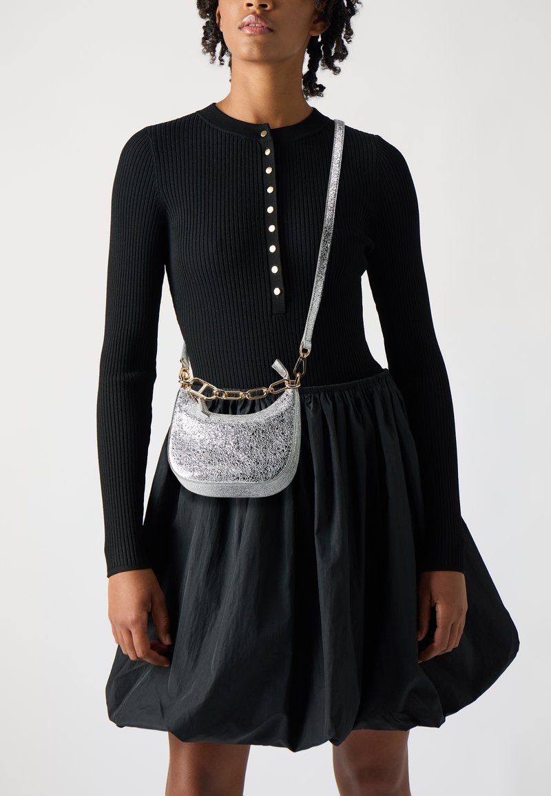 Haut noir à rayures avec des boutons argentés, associé à une jupe noire volumineuse. Un petit sac en bandoulière métallique argenté avec une chaîne est présenté.