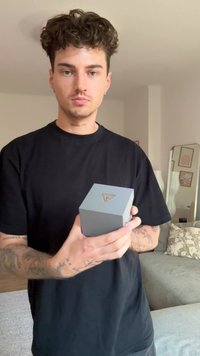 Pelēks kastīte ar trīsstūra logotipu augšā, ko tur cilvēks melnā kreklā. Kastei ir gluda virsma un minimālistisks dizains.