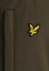 Il tessuto verde oliva presenta una trama liscia con cuciture in evidenza. Un logo di un'aquila ricamato di colore giallo è visibile, aggiungendo dettaglio al design.