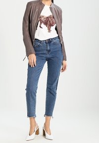 Ruskea nahkainen biker-takki, jossa on resorutetut olkapäät, valkoinen t-paita, jossa on leijonakuva, siniset korkeavyötäröiset farkut ja valkoiset, lohkoluvulla varustetut korot.