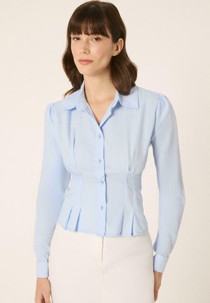 Femme portant une blouse cintrée bleu clair à boutons, manches longues et col, associée à un pantalon blanc sur un fond uni.