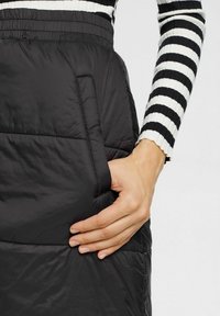 Gonna a sacco nera con una texture liscia e una vita elasticizzata. Presenta una tasca laterale e delle cuciture decorative per dare forma e struttura.