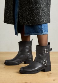 Black leather ankle boots with a rounded toe, adjustable straps, silver buckles, and a chunky heel, set against a wooden floor.