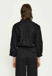 Persoană cu păr brunet de lungime medie, îmbrăcată cu o cămașă neagră cu mâneci lungi și largi, și pantaloni negri, stând cu spatele, pe un fundal simplu.