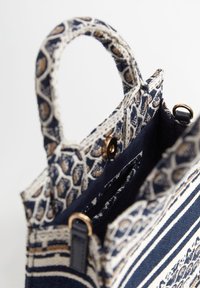 Pieni, jäsennelty laukku, jossa on kudottu kangas tummansinisin, kerma- ja kultavärisin kuosein. Varustettu kahdella matalalla kahvalla ja magneettisella napilla.