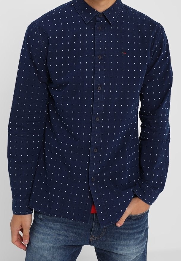 Chemise boutonnée bleu marine à pois blancs, avec un col, des manches longues et un petit logo sur la poitrine. Tissu texturé.