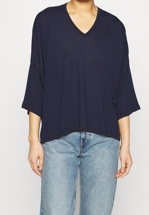 Henkilö, joka käyttää löysää laivastonsinistä V-aukkokauluksellista yläosaa, jossa on kolmen neljänneksen hihat, ja vaaleansinisiä farkkuja yksiväristä taustaa vasten.