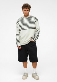 Lichtgrijs, langemouwen shirt met zwarte horizontale strepen, gecombineerd met zwarte shorts. De outfit wordt compleet gemaakt met grijze sneakers en witte sokken.