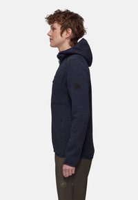 Giacca in pile blu navy con cappuccio, tessuto testurizzato, tasca frontale con zip e patch con logo sulla manica sinistra. Indossata con pantaloni verde oliva.