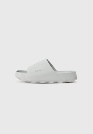 Witte Nike slidesandaal met een dikke band en een gestructureerde zool, vanaf de zijkant getoond op een effen grijze achtergrond.