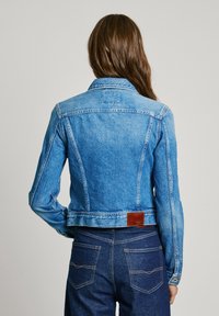 Light blue denim jacket featuring a fitted design, prominent stitching, and a brown leather patch at the back. Long sleeves and classic collar.