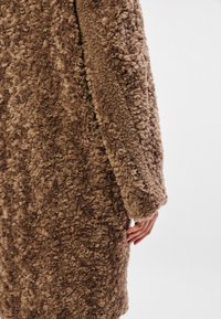 Καφέ τεχνητό παλτό φτιαγμένο από γούνα με υφή που θυμίζει μαλλί, σκούρα και φαρδιά μανίκια και χαλαρή εφαρμογή. Χέρι που στηρίζεται απαλά στο ύφασμα.