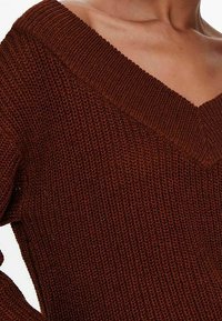 Pull marron tricoté avec un large col en V, présentant un tissu texturé et des détails à ourlet côtelé. Vue rapprochée du décolleté et de la zone des épaules.