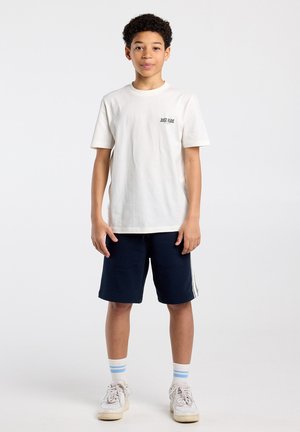 Jongen staand, draagt een wit "Just Ride" T-shirt, marineblauwe korte broek met witte strepen, witte sokken met blauwe strepen en witte sneakers.