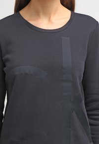 Mörkgrå sweatshirt med en texturerad, överdimensionerad logotyp på framsidan. Den har en rund halsringning och en mjuk tygtextur.