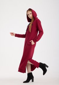 Lange bordeauxrode hoodie-jurk met zijslitsen, zak en een capuchon met koord; gecombineerd met zwarte enkellaarsjes. Zachte stof met een ontspannen pasvorm.
