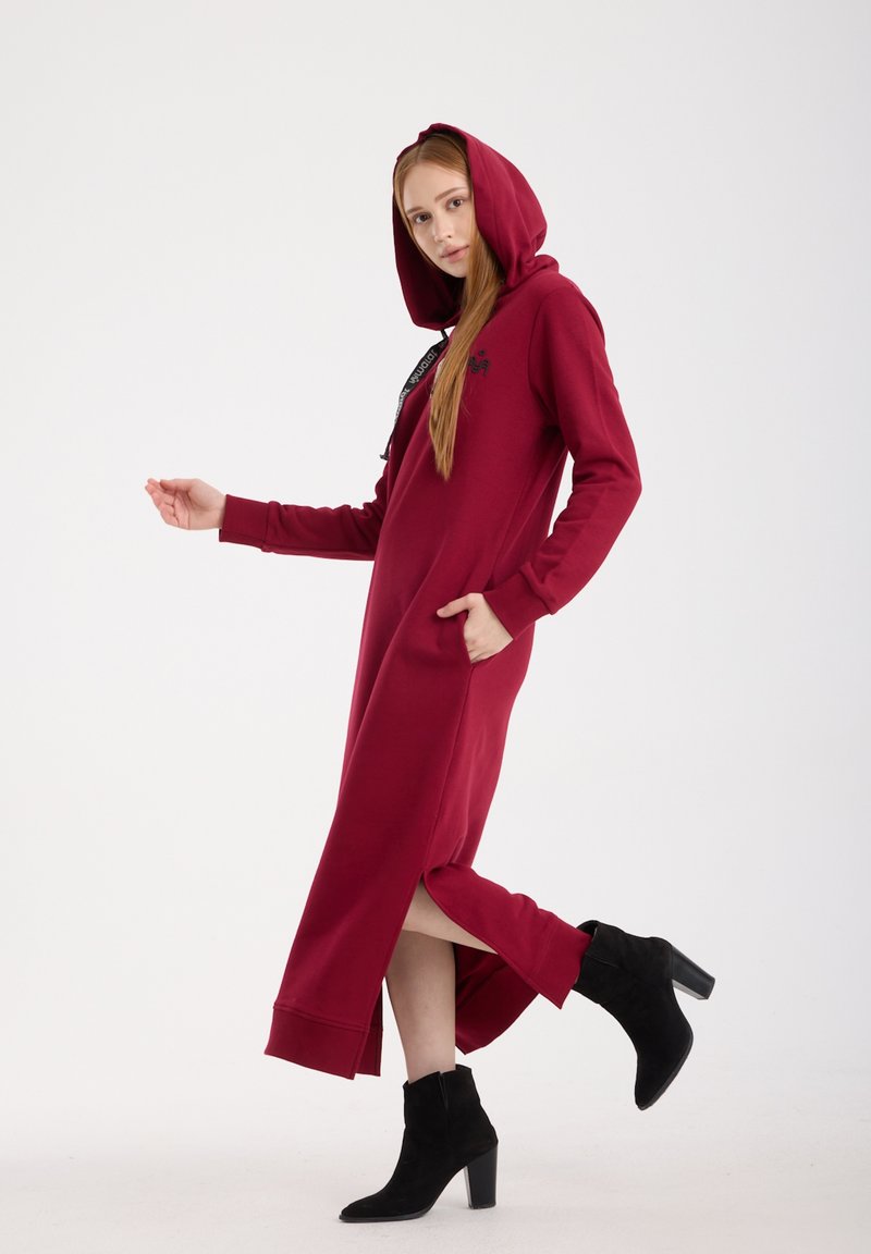 Lange bordeauxrode hoodie-jurk met zijslitsen, zak en een capuchon met koord; gecombineerd met zwarte enkellaarsjes. Zachte stof met een ontspannen pasvorm.