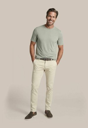 Man staat met handen in de zakken, draagt een lichtgroen t-shirt, beige broek, bruine riem en bruine slip-on schoenen tegen een effen achtergrond.