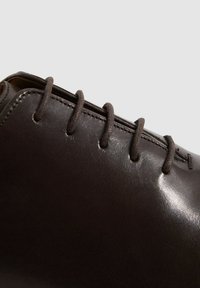 Braunes Leder-Schuh mit glatter Textur, ausgestattet mit sechs Ösen und runden Schnürsenkeln. Das Design ist elegant und betont eine glänzende Oberfläche.