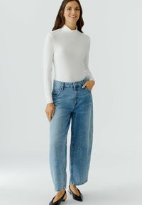 Weißes langärmliges Rollkragenoberteil, kombiniert mit locker sitzenden hellblauen Jeans aus Denim mit sichtbaren Nähten und einer entspannten Silhouette. Schwarze Schuhe.