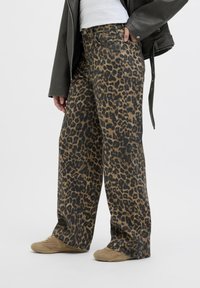 Persona que viste pantalones anchos con estampado de leopardo, zapatillas marrones claras, camisa blanca y chaqueta de cuero negra con la mano en el bolsillo sobre un fondo liso.