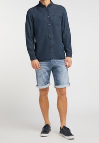 Ciemnoniebieska koszula na guziki z kieszenią, zestawiona z jasnobłękitnymi szortami jeansowymi podwiniętymi na brzegach oraz ciemnymi trampkami.