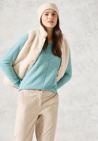 Beige Fleece-Weste über einem hellblauen gerippten Langarmshirt und einer cremefarbenen Strickmütze, kombiniert mit hellen khakifarbenen Hosen, vor einem strukturierten Hintergrund.