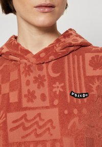 Sudadera con capucha en tejido de toalla texturizado, color óxido, que presenta varios patrones en relieve y un parche negro con el logo "VOLCOM".