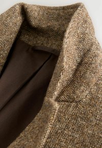 Bruine tweed blazer met textuurstof, brede revers en een bruine voering. Close-upweergave toont de kraag en stikseldetails.