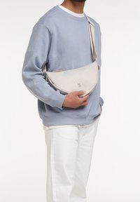Sac bandoulière beige avec une texture lisse, présentant un design simple et une bandoulière, tenu par une personne portant un sweat-shirt bleu clair.