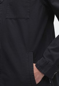 Gros plan d'une personne portant une veste noire à manches longues avec une main partiellement dans une poche latérale.