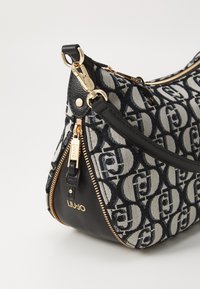 Borsa a spalla nera e beige con tessuto stampato, dettagli in oro, zip e una base in pelle a contrasto. Logo visibile sul lato.