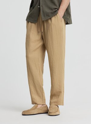 Uomo che indossa pantaloni beige larghi con coulisse e mocassini in camoscio abbinati, mani in tasca, su uno sfondo chiaro e uniforme.