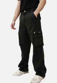 Pantaloni cargo neri in cotone, con più tasche, vestibilità relax e chiusura con bottone. Indossati con scarpe sportive bianche.