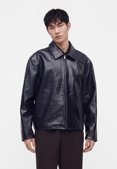 Jeune homme portant une veste en cuir noire zippée avec un large col, se tenant devant un fond gris clair uni.