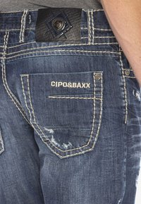 Jeans aus Denim mit Distressed-Details, dunkelblauer Farbe, kontrastierenden weißen Nähten und einem strukturierten Lederpatch am Bund mit der Aufschrift CIPO&BAXX.