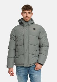 Homme portant une veste d'hiver grise rembourrée avec capuche et poches latérales, associée à un jean bleu, sur fond blanc uni.