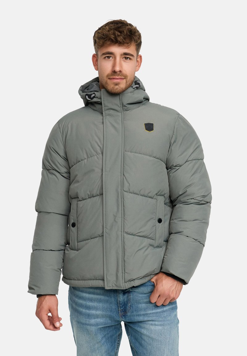 Mężczyzna w szarej ocieplanej kurtce zimowej z kapturem i bocznymi kieszeniami, w połączeniu z niebieskimi dżinsami, na tle jednolitego białego tła.