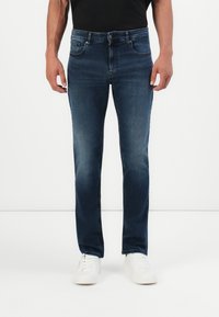 Mees, kes kannab tumedat sinist kitsast teksapükste ja valgete tossudega, seisab ühtlase valge tausta ees.