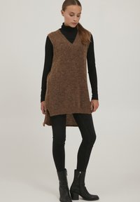 Brun, strikket lang vest med V-hals, sideslids og løs pasform, parret med en sort rullekrave og sorte skinny jeans samt støvler.
