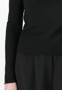 Cămașă neagră cu mâneci lungi, cu un tiv ondulat. Confecționată dintr-un material moale, având un design ajustat cu detalii subtile de textură.