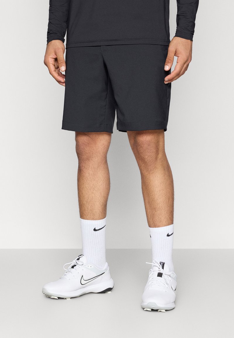 Schwarze Sportshorts mit glatter Textur, lockerer Passform und seitlichen Taschen. Getragen mit weißen Nike-Socken und passenden Turnschuhen.