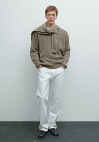 Gebreide trui in taupe met ribbelstructuur en ronde hals, gelaagd met een bijpassende sjaal; gecombineerd met witte wijde broek en zwarte schoenen.